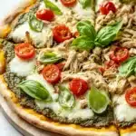 Pesto chicken pizza - the image is of a freshly baked pizza on a wooden cutting board. the pizza has a golden brown crust and is topped with shredded chicken, sliced cherry tomatoes, and fresh basil leaves. the chicken appears to be cooked and is arranged in a circular pattern on top of the pizza. the basil leaves are scattered around the pizza, adding a pop of green color to the dish. the background is a white marble countertop.