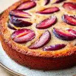 Plum cake allo yogurt giallo zafferano - the image is of a round cake on a white plate. the cake appears to be a cherry tart, with a golden brown crust and a flaky, buttery interior. on top of the cake, there are sliced plums arranged in a circular pattern. the plums are dark red in color and have a glossy sheen. the plate is sitting on a wooden table, and the background is blurred, making the cake the focal point of the image.