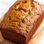 Pumpkin apple zucchini bread - the image shows a freshly baked loaf of bread on a wooden cutting board. the bread appears to be golden brown in color and has a crumbly texture. it is topped with sliced zucchini, which are visible in the center of the loaf. the cutting board is placed on a white marble countertop. the background is blurred, making the bread the focal point of the image.