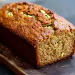 Pumpkin apple zucchini bread - the image shows a freshly baked loaf of zucchini bread on a wooden cutting board. the bread is golden brown in color and has a crumbly texture on top. it appears to be moist and fluffy, with small zucchinis scattered throughout. the cutting board is placed on a dark blue countertop, and the background is blurred, making the bread the focal point of the image.