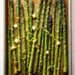 Roasted lemon honey garlic asparagus - the image shows a baking tray with cooked asparagus spears on it. the tray is rectangular in shape and has a light brown color. the spears are arranged in a single layer on the tray, with some of them overlapping each other. they are coated in a dark brown sauce, which appears to be seasoned with herbs and spices. there are also small pieces of garlic scattered throughout the tray. the background is a white wooden surface.