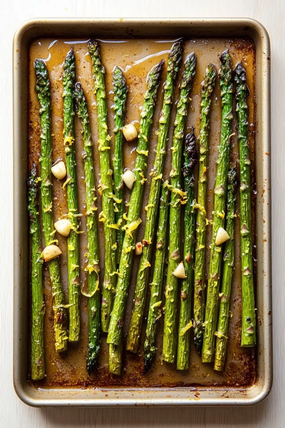 Roasted lemon honey garlic asparagus - the image shows a baking tray with cooked asparagus spears on it. the tray is rectangular in shape and has a light brown color. the spears are arranged in a single layer on the tray, with some of them overlapping each other. they are coated in a dark brown sauce, which appears to be seasoned with herbs and spices. there are also small pieces of garlic scattered throughout the tray. the background is a white wooden surface.