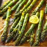 Roasted lemon honey garlic asparagus - the image shows a tray of cooked asparagus spears on a wooden table. the spears are arranged in a single layer on the tray, with some of them overlapping each other. they are covered in a dark sauce, which appears to be drizzled over the top of the spears. there are also a few slices of lemon scattered around the tray. the background is blurred, but it seems to be a kitchen countertop. the overall mood of the image is fresh and appetizing.