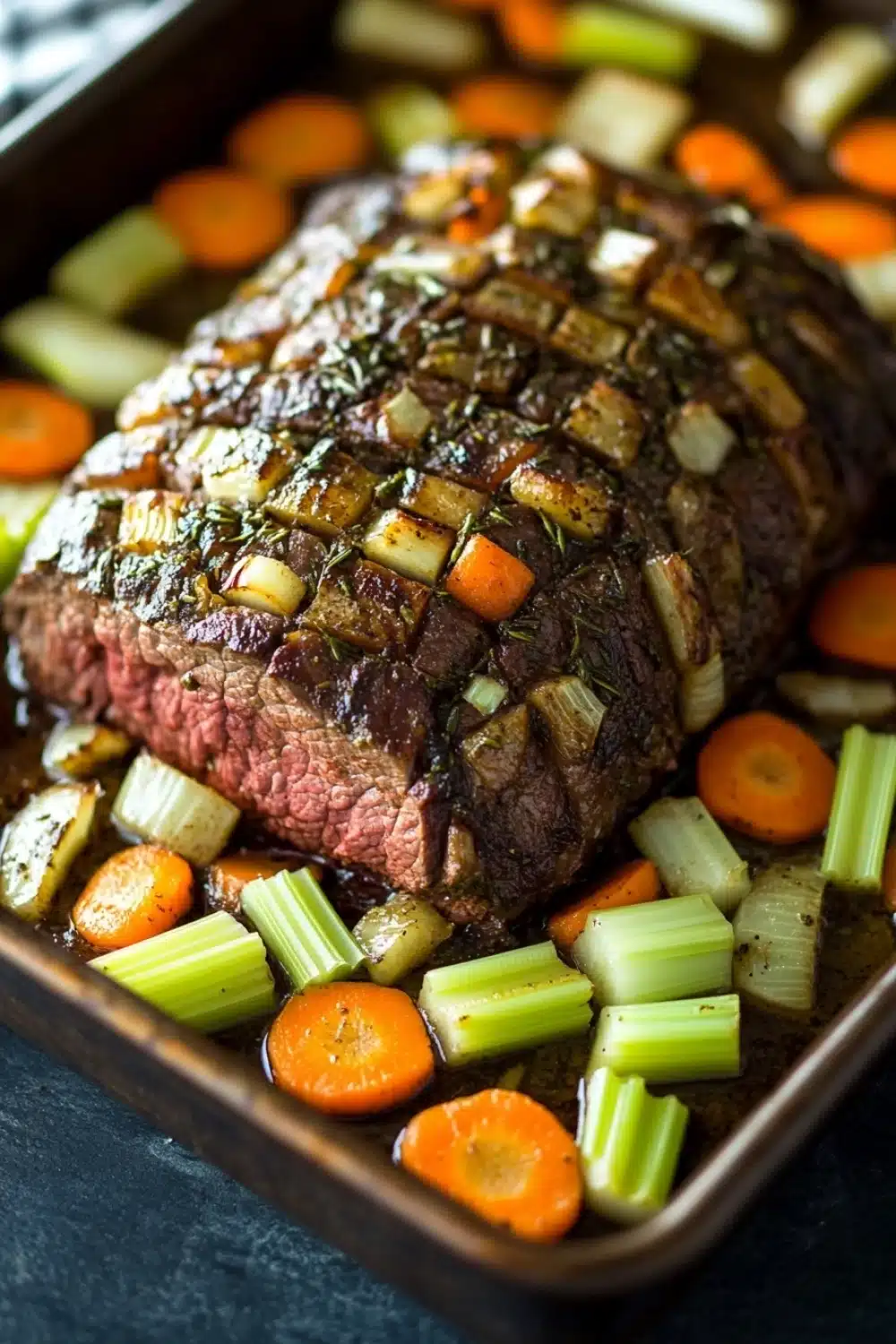 Rosemary garlic roast beef slow cooker - the image shows a large piece of cooked steak on a baking tray. the steak appears to be medium-rare and has a pinkish-brown crust on top. it is garnished with herbs and spices, including rosemary and thyme. the vegetables on the tray include carrots, celery, and onions. the tray is lined with parchment paper and there is a blue and white striped napkin in the background. the overall presentation of the dish is elegant and appetizing.