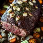 Rosemary garlic roast beef slow cooker - the image shows a large piece of cooked steak on a black baking tray. the steak appears to be medium-rare and has a dark brown crust on top. it is garnished with chopped onions and rosemary sprigs. there are also small pieces of carrots and celery scattered around the steak. the tray is sitting on a wooden table.