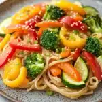 Sauteed vegetables with noodles - the image is a close-up of a plate of noodles with vegetables. the noodles are light brown and appear to be stir-fried with sesame seeds. the vegetables are mixed in with the noodles, including broccoli, red and yellow bell peppers, and sliced cucumbers. the colors of the vegetables are vibrant and the dish looks fresh and appetizing. the plate is made of a dark brown ceramic with a speckled pattern. the background is white, making the colors of food stand out.