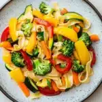 Sauteed vegetables with noodles - the image is a close-up of a plate of pasta salad. the plate is round and has a light blue rim with a speckled pattern. the pasta is fettuccine alfredo and is mixed with various vegetables such as bell peppers, broccoli, carrots, and zucchini. the vegetables are bright yellow and red in color, and they are arranged in a colorful and appetizing manner. the dish appears to be freshly cooked and ready to eat.