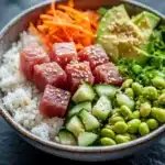 Seared tuna poke bowl - the image is a close-up of a bowl of food. the bowl is made of ceramic and is placed on a dark grey surface. the food in the bowl appears to be a salad with a variety of ingredients. on the left side of the bowl, there is a bed of white rice, topped with sliced carrots, sliced cucumbers, and sliced avocado. the vegetables are arranged in a colorful and appetizing manner. the colors of the ingredients are vibrant and the dish looks fresh and fresh. the background is blurred, making the bowl the focal point of the image.