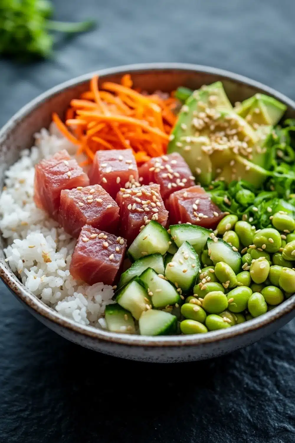 Seared tuna poke bowl - the image is a close-up of a bowl of food. the bowl is made of ceramic and is placed on a dark grey surface. the food in the bowl appears to be a salad with a variety of ingredients. on the left side of the bowl, there is a bed of white rice, topped with sliced carrots, sliced cucumbers, and sliced avocado. the vegetables are arranged in a colorful and appetizing manner. the colors of the ingredients are vibrant and the dish looks fresh and fresh. the background is blurred, making the bowl the focal point of the image.