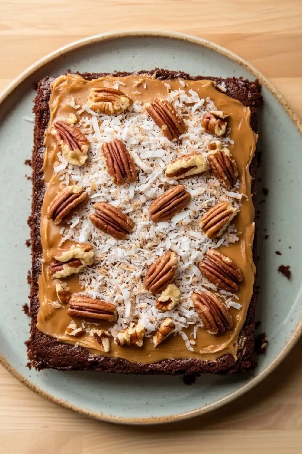 Sheet cake german chocolate - the image is of a square cake on a blue plate. the cake appears to be a chocolate brownie with a layer of caramel on top. the caramel is drizzled over the top of the cake, creating a smooth and creamy texture. there are also several pecans scattered on top, adding a crunchy texture to the cake. the plate is sitting on a wooden table.