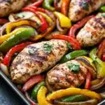 Sheet pan chicken breast with fajita veggies - the image shows a baking tray filled with grilled chicken and colorful bell peppers. the chicken is golden brown and appears to be seasoned with herbs and spices. the bell peppers are bright red, yellow, and green, and they are arranged in a circular pattern around the chicken. the tray is lined with parchment paper and is placed on a black countertop. the overall presentation of the dish is colorful and appetizing.