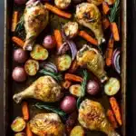 Sheet pan chicken legs and vegetables - the image shows a rectangular baking tray filled with roasted chicken and vegetables. the chicken is golden brown and appears to be seasoned with herbs and spices. the vegetables include carrots, potatoes, onions, and rosemary sprigs. the tray is lined with parchment paper and is placed on a wooden table. the overall presentation of the dish is colorful and appetizing.