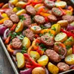 Sheet pan dinner sausage and potatoes - the image shows a rectangular baking tray filled with a colorful dish. the dish appears to be a mix of diced potatoes, red and yellow bell peppers, onions, and sausage. the potatoes are cut into small cubes and are arranged in a pile on top of each other. the sausage is cooked to a golden brown color and is garnished with fresh herbs. the vegetables are mixed together with the sausage and onions, creating a colorful and appetizing presentation. the tray is sitting on a wooden table with a sprig of parsley on the side.