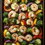 Sheet pan dinner with shrimp and vegetables - the image shows a rectangular baking tray filled with cooked shrimp and vegetables. the shrimp are pink and appear to be seasoned with herbs and spices. the vegetables include broccoli, zucchini, red onions, and yellow bell peppers. the tray is placed on a wooden table with a gray cloth napkin on the side. the background is dark, making the colors of the vegetables stand out.