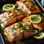 Sheet pan garlic lemon butter salmon - the image shows four pieces of salmon fillets on a baking tray. the salmon is cooked to a golden brown color and is garnished with fresh herbs and lemon wedges. the tray is placed on a dark wooden table with a few sprigs of parsley scattered around it. the background is blurred, but it appears to be a kitchen countertop. the overall mood of the image is fresh and appetizing.