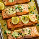 Sheet pan garlic lemon butter salmon - the image shows three pieces of salmon on a baking tray. the salmon is cooked to a golden brown color and is garnished with lemon wedges and chopped parsley. the tray is lined with parchment paper and there is a white napkin on the side. the background is blurred, but it appears to be a wooden table.