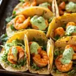 Sheet pan spicy shrimp tacos with jalapeno lime crema - the image shows a tray of shrimp tacos on a wooden table. the tacos are made with a tortilla shell and are filled with shrimp, lettuce, and a creamy green sauce. the shrimp are cooked medium-sized and appear to be seasoned with herbs and spices. the tray is lined with cilantro leaves, adding a pop of green color to the dish. the background is blurred, but it appears to be a kitchen countertop.