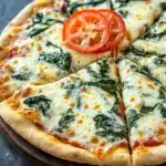 Sheet pan spinach dip pizza - the image is of a freshly baked pizza on a wooden cutting board. the pizza has a golden brown crust and is topped with melted cheese and fresh spinach leaves. there are two slices of tomato on top of the cheese, one on each side of the pizza. the background is a dark grey countertop.