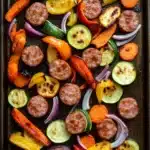 Sheet pan vegetables and sausage - the image shows a baking tray lined with parchment paper. on the tray, there are various types of vegetables, including sliced zucchini, red onions, and yellow bell peppers. the vegetables are arranged in a circular pattern, with some overlapping each other. the tray appears to be freshly baked and ready to be served. the background is dark, making the colors of the vegetables stand out.