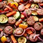 Sheet pan vegetables and sausage - the image shows a tray of roasted vegetables on a wooden table. the vegetables are arranged in a circular pattern on the tray, with some overlapping each other. there are small pieces of sausage, zucchini, carrots, and bell peppers scattered throughout the tray. the sausage is golden brown and appears to be seasoned with herbs and spices. the bell peppers are bright orange and yellow, and the onions are red. the tray is lined with parchment paper, and there is a sprinkle of herbs on top. the overall appearance of the dish is colorful and appetizing.