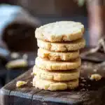 Shortbread cookie illustration - the image shows a stack of freshly baked cookies on a wooden cutting board. the cookies are golden brown in color and appear to be soft and chewy. they are stacked on top of each other, with the top cookie slightly overlapping the bottom one. the cutting board is placed on a rustic wooden table, with a blurred background of greenery and a white cloth napkin. the overall mood of the image is warm and inviting.