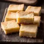 Shortbread cookie squares - the image shows a wooden cutting board with several square-shaped bars stacked on top of each other. the bars appear to be freshly baked and have a golden-brown color with a crumbly texture. they are arranged in a neat row on the board, with some overlapping each other, and there are a few more bars visible in the background. the cutting board is placed on a dark wooden table, and the background is blurred, making the bars the focal point of the image.