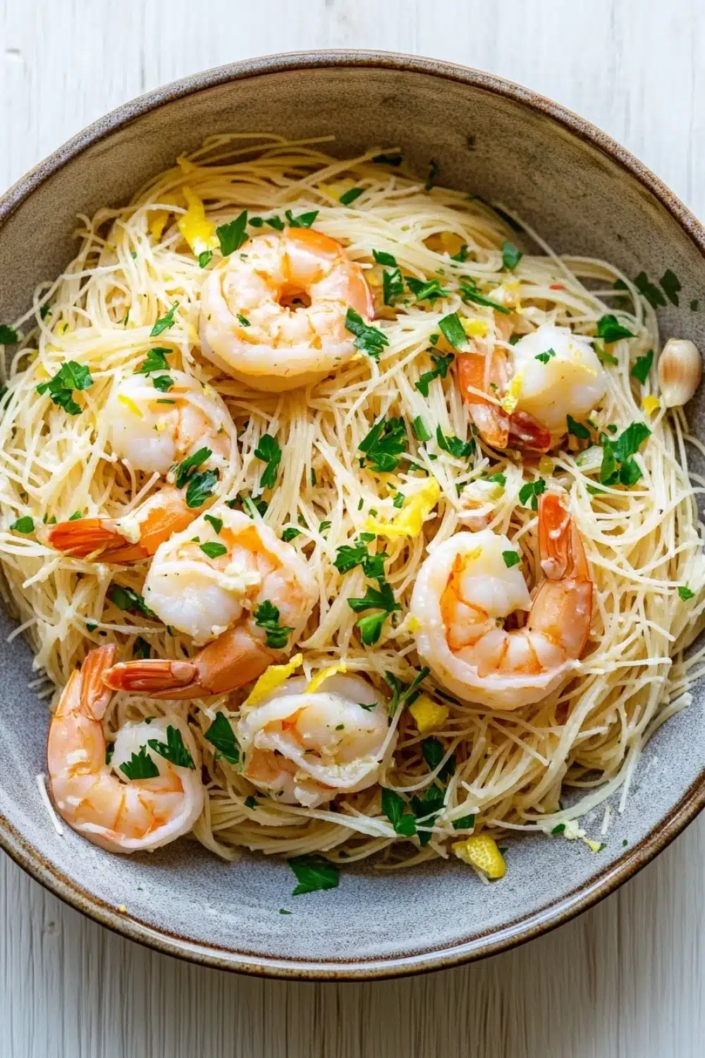 Shrimp scampi angel hair pasta - the image is a close-up of a bowl of spaghetti with shrimp and lemon zest. the spaghetti is cooked al dente and is garnished with chopped parsley and garlic cloves. the shrimp are large and pink, and they are arranged in a circular pattern on top of the spaghetti. the dish is served in a light brown bowl with a white rim. the background is a white wooden table.