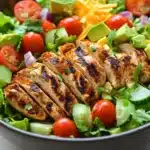 Siracha lime chicken chopped salad - the image is a close-up of a large bowl of salad. the salad is made up of various types of vegetables, including lettuce, cherry tomatoes, cucumbers, avocado, and shredded cheese. on top of the salad, there are grilled chicken pieces that have been grilled to a golden brown color. the chicken appears to be seasoned with herbs and spices, giving it a savory flavor. the bowl is sitting on a wooden table, and the background is blurred, making the salad the focal point of the image.