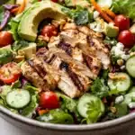 Siracha lime chicken chopped salad - the image is a close-up of a large bowl of salad. the salad is made up of various greens, including lettuce, cherry tomatoes, cucumbers, and red cabbage. on top of the salad, there are slices of grilled chicken, sliced avocado, and chopped nuts. the chicken appears to be seasoned with herbs and spices, giving it a savory flavor. the bowl is sitting on a white wooden table.
