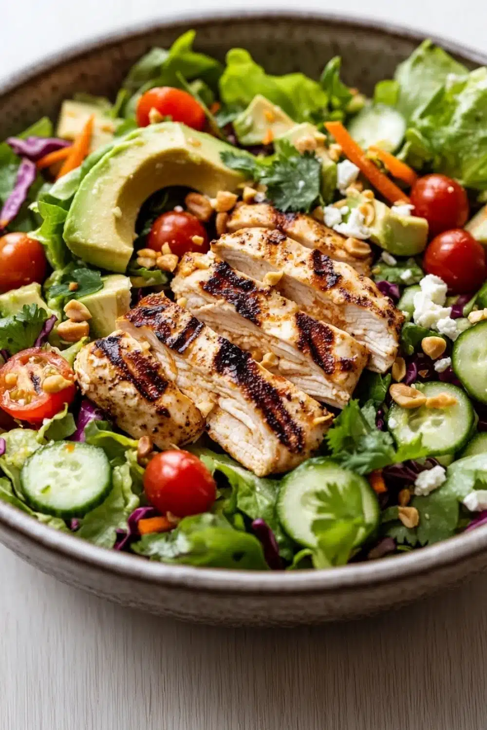 Siracha lime chicken chopped salad - the image is a close-up of a large bowl of salad. the salad is made up of various greens, including lettuce, cherry tomatoes, cucumbers, and red cabbage. on top of the salad, there are slices of grilled chicken, sliced avocado, and chopped nuts. the chicken appears to be seasoned with herbs and spices, giving it a savory flavor. the bowl is sitting on a white wooden table.