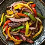 Slow cooker beef fajitas slow cooker - the image is a close-up of a plate of food. the plate is made of ceramic and has a dark brown color. the food appears to be a stir-fry dish with sliced beef and colorful bell peppers. the beef is cooked medium-rare and the peppers are bright red, yellow, and green. the peppers are arranged in a circular pattern around the beef, with some overlapping each other. the dish is garnished with a sprinkle of herbs and spices. the background is a wooden table with a rustic texture.