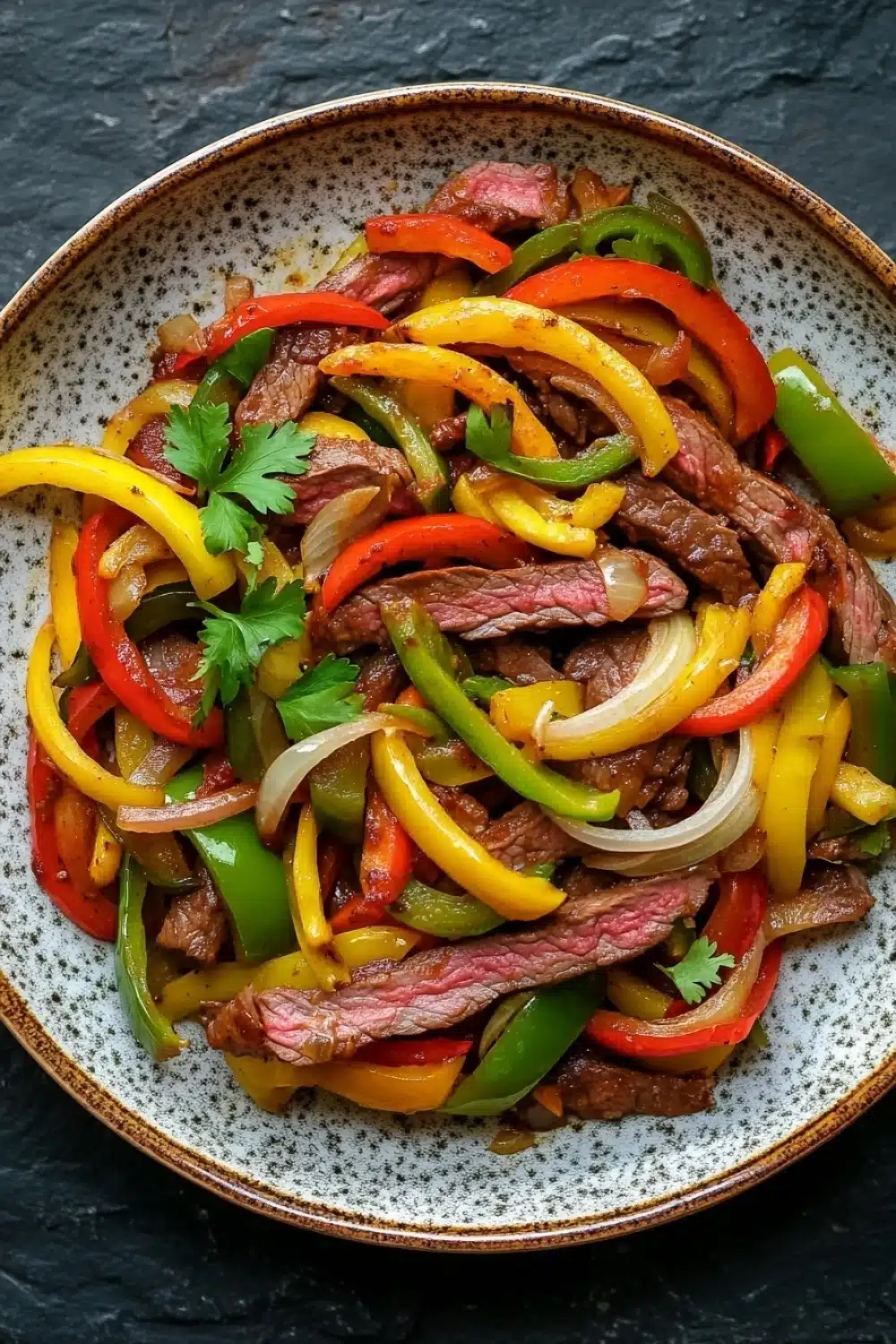 Slow cooker beef fajitas slow cooker - the image is a close-up of a plate of food. the plate is round and has a speckled pattern around the edges. the food appears to be a stir-fry with sliced beef, red and yellow bell peppers, and sliced onions. the beef is cooked medium-rare and is arranged in a circular pattern on top of the peppers. the peppers are bright red, yellow, and green, and the onions are sliced into thin strips. there is a sprig of cilantro on top, adding a pop of green to the dish. the background is a dark grey textured surface.