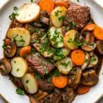 Slow cooker beef hotpot - the image is a close-up of a dish in a white bowl with a brown rim. the dish appears to be a beef stew with chunks of beef, potatoes, carrots, and mushrooms. the beef is cooked medium-rare and is garnished with chopped parsley. the potatoes are cut into small cubes and are arranged in a pile on top of the beef. the carrots are bright orange and the mushrooms are sliced into thin slices. the broth is a dark brown color and looks thick and flavorful. the bowl is sitting on a white marble countertop with a few sprigs of parsley scattered around.