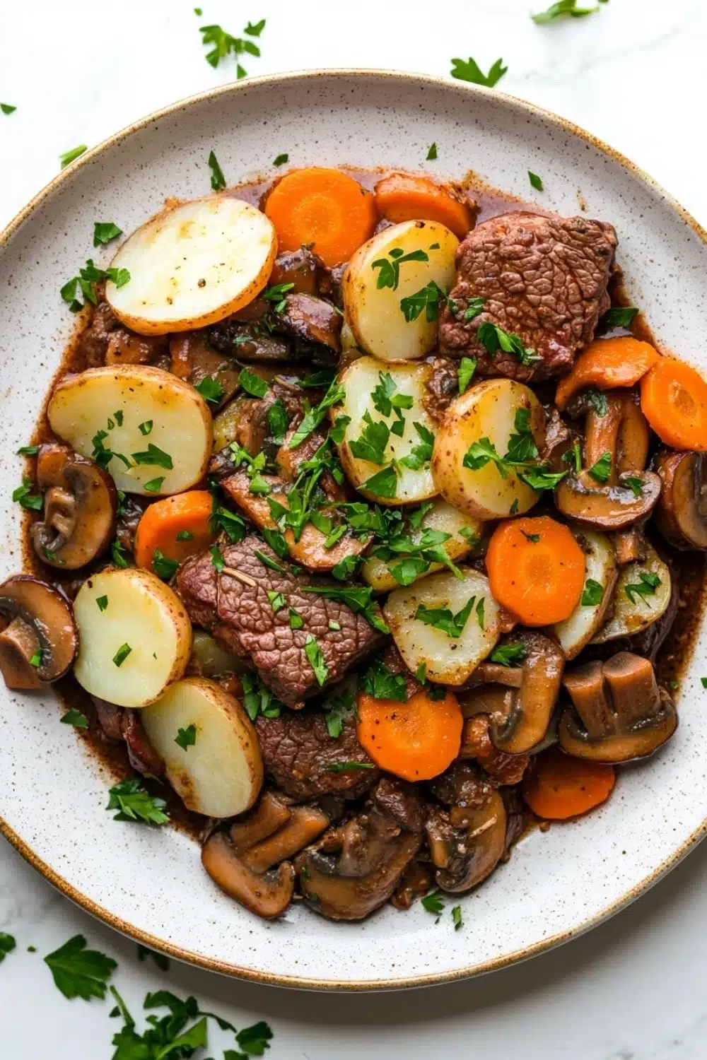Slow cooker beef hotpot - the image is a close-up of a dish in a white bowl with a brown rim. the dish appears to be a beef stew with chunks of beef, potatoes, carrots, and mushrooms. the beef is cooked medium-rare and is garnished with chopped parsley. the potatoes are cut into small cubes and are arranged in a pile on top of the beef. the carrots are bright orange and the mushrooms are sliced into thin slices. the broth is a dark brown color and looks thick and flavorful. the bowl is sitting on a white marble countertop with a few sprigs of parsley scattered around.