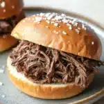Slow cooker beef on weck - the image shows two pulled pork sandwiches on a blue and white plate. the sandwiches are made with a sesame seed bun and are topped with shredded beef. the beef appears to be cooked and has a dark brown color. the plate is sitting on a wooden table with a blurred background.