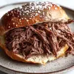 Slow cooker beef on weck - the image shows a plate with a sandwich on it. the sandwich is made with a toasted bun and is covered in shredded beef. the beef appears to be cooked and has a dark brown color. the plate is white with a speckled pattern around the edges. the background is blurred, but it seems to be a kitchen countertop.