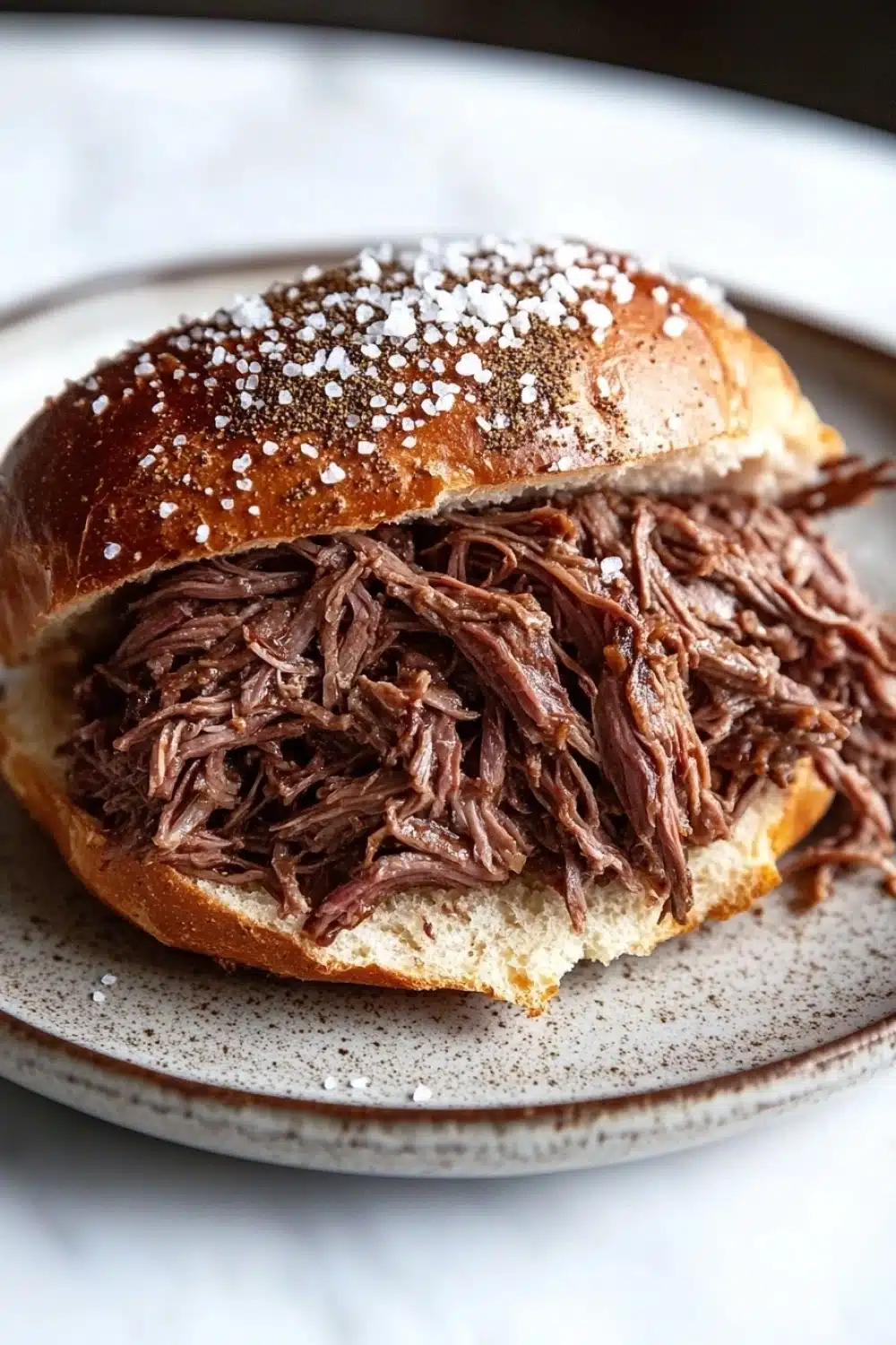 Slow cooker beef on weck - the image shows a plate with a sandwich on it. the sandwich is made with a toasted bun and is covered in shredded beef. the beef appears to be cooked and has a dark brown color. the plate is white with a speckled pattern around the edges. the background is blurred, but it seems to be a kitchen countertop.
