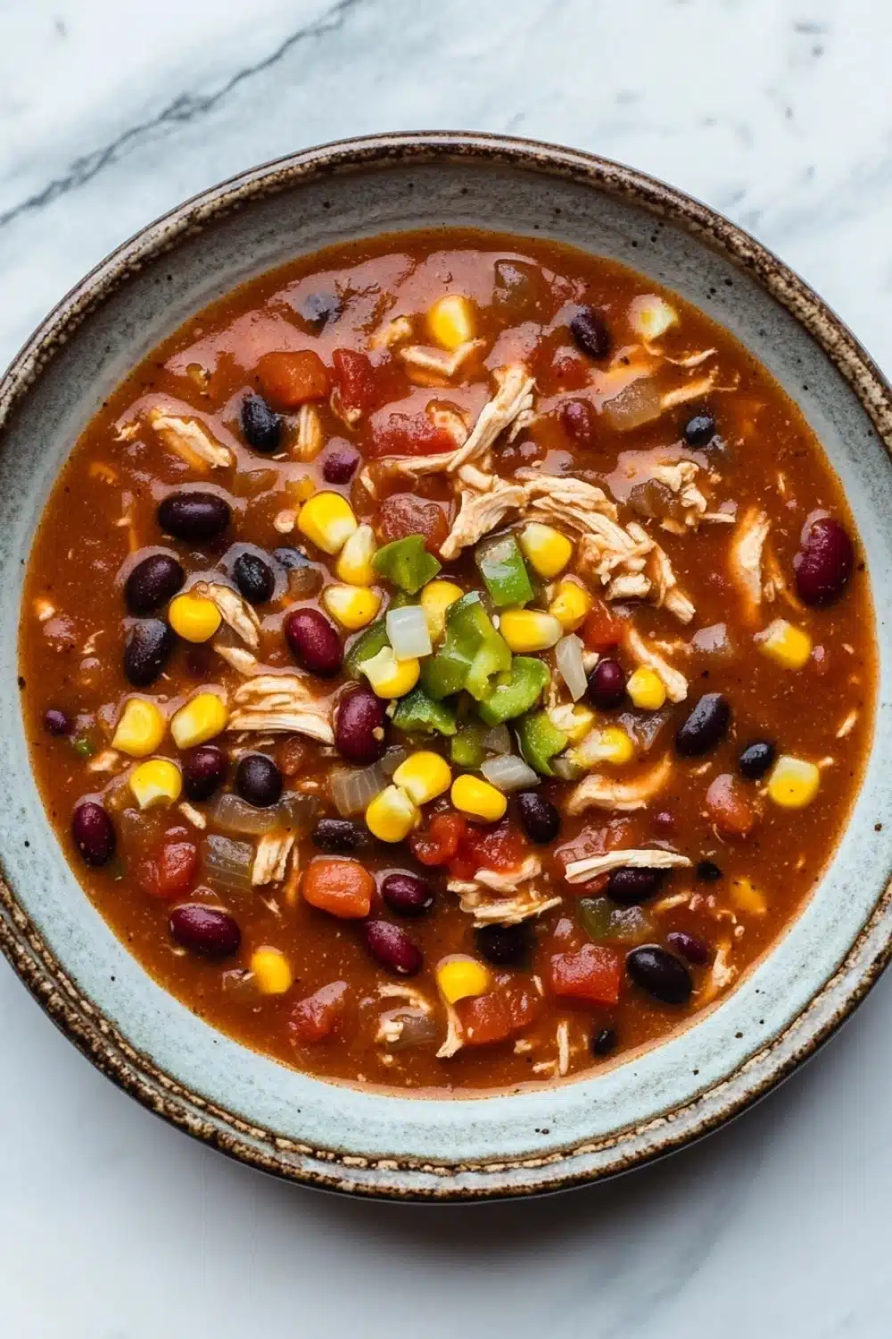 Slow cooker chicken chili - the image is a close-up of a bowl of chili. the chili is a rich orange color and appears to be thick and hearty. it is filled with chunks of chicken, diced tomatoes, black beans, corn kernels, and green bell peppers. the bowl is made of ceramic and has a light blue rim. the background is a white marble countertop.