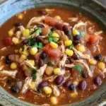 Slow cooker chicken chili - the image is a close-up of a bowl of chili. the chili is a rich orange color and appears to be made with chunks of chicken, black beans, corn kernels, and other vegetables. the bowl is made of ceramic and is sitting on a wooden table. there are a few sprigs of parsley scattered around the bowl. the background is blurred, but it seems to be a rustic kitchen countertop.