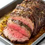 Slow cooker deli roast beef - the image shows a large piece of cooked steak on a baking tray. the steak appears to be medium-rare and has a pinkish-red color. it is sliced into thin strips and is covered in a black pepper sauce. the sauce is drizzled over the top of the steak, giving it a glossy sheen. the tray is lined with parchment paper and there is a white cloth napkin on the side. the background is a plain white countertop.