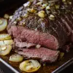 Slow cooker deli roast beef - the image shows a large piece of cooked steak on a black baking tray. the steak appears to be medium-rare and has a pinkish-brown crust on top. it is garnished with chopped herbs and sliced onions. the tray is sitting on a wooden table with a blue and white striped napkin underneath. the overall presentation of the dish is elegant and appetizing.