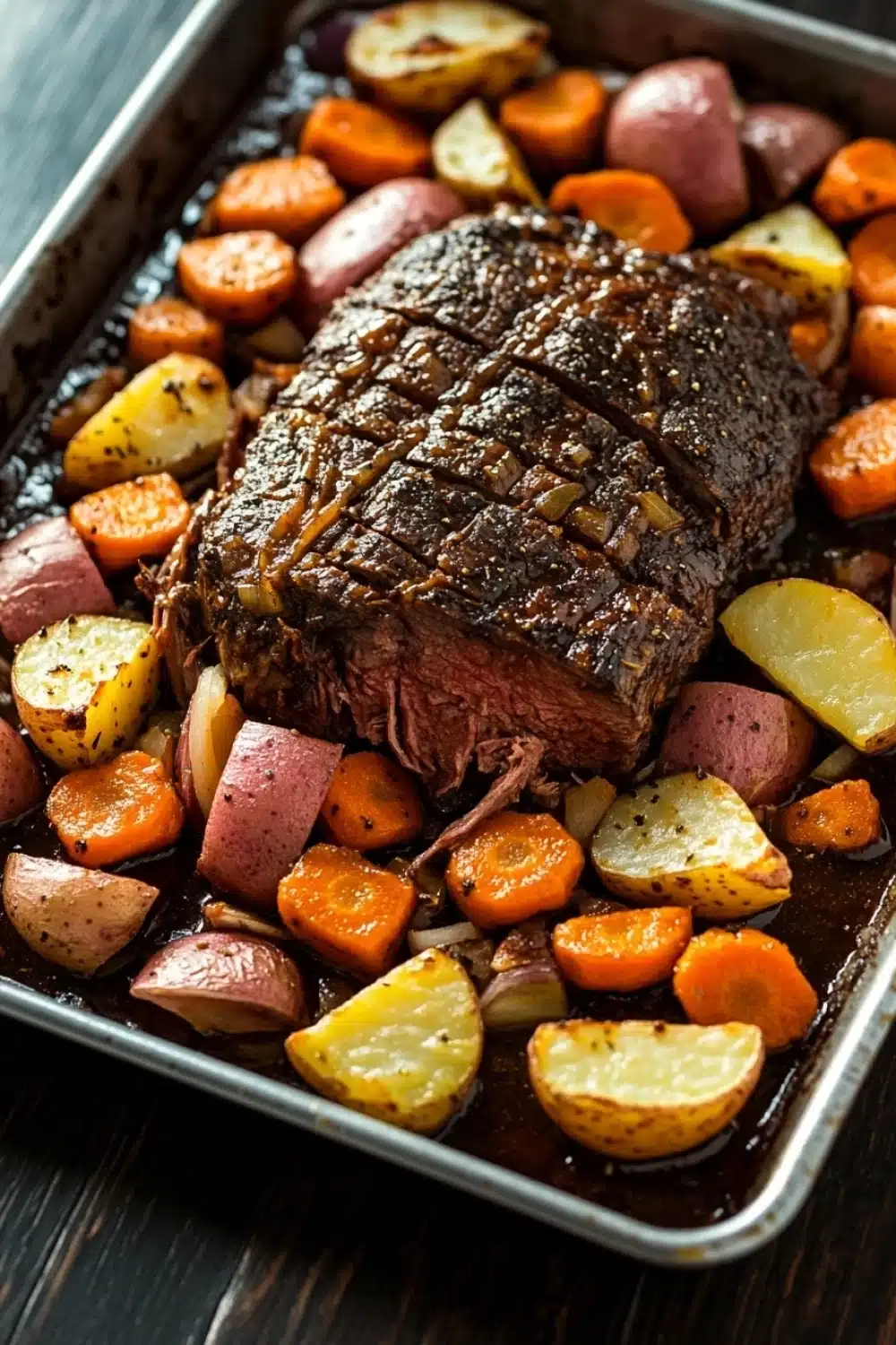 Slow cooker pot roast and vegetables - the image shows a rectangular baking tray with a large piece of roast beef on it. the roast beef is cooked medium-rare and has a golden brown crust on top. it is surrounded by roasted vegetables, including carrots, potatoes, and onions. the vegetables are arranged in a pile on the tray, with some overlapping each other. the tray is placed on a wooden table, and the background is blurred, making the focus of the image the roast beef and vegetables.