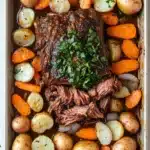 Slow cooker pot roast beef - the image shows a rectangular baking dish filled with a roast beef dish. the roast beef is cooked medium-rare and is garnished with a sprig of fresh parsley. the dish is served on a bed of roasted potatoes and carrots. the potatoes are golden brown and appear to be seasoned with herbs and spices. the carrots are bright orange and look ripe and juicy. the onions are white and are scattered throughout the dish, adding a pop of color to the overall appearance. the tray is lined with parchment paper and is sitting on a white countertop.