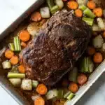 Slow cooker roast beef from frozen - the image shows a rectangular baking dish with a large piece of cooked meat in it. the meat appears to be a roast beef, with a dark brown crust on top. it is surrounded by a bed of diced carrots, celery, and onions. the dish is sitting on a white marble countertop. the vegetables are arranged in a way that they are overlapping each other, creating a colorful and appetizing display.
