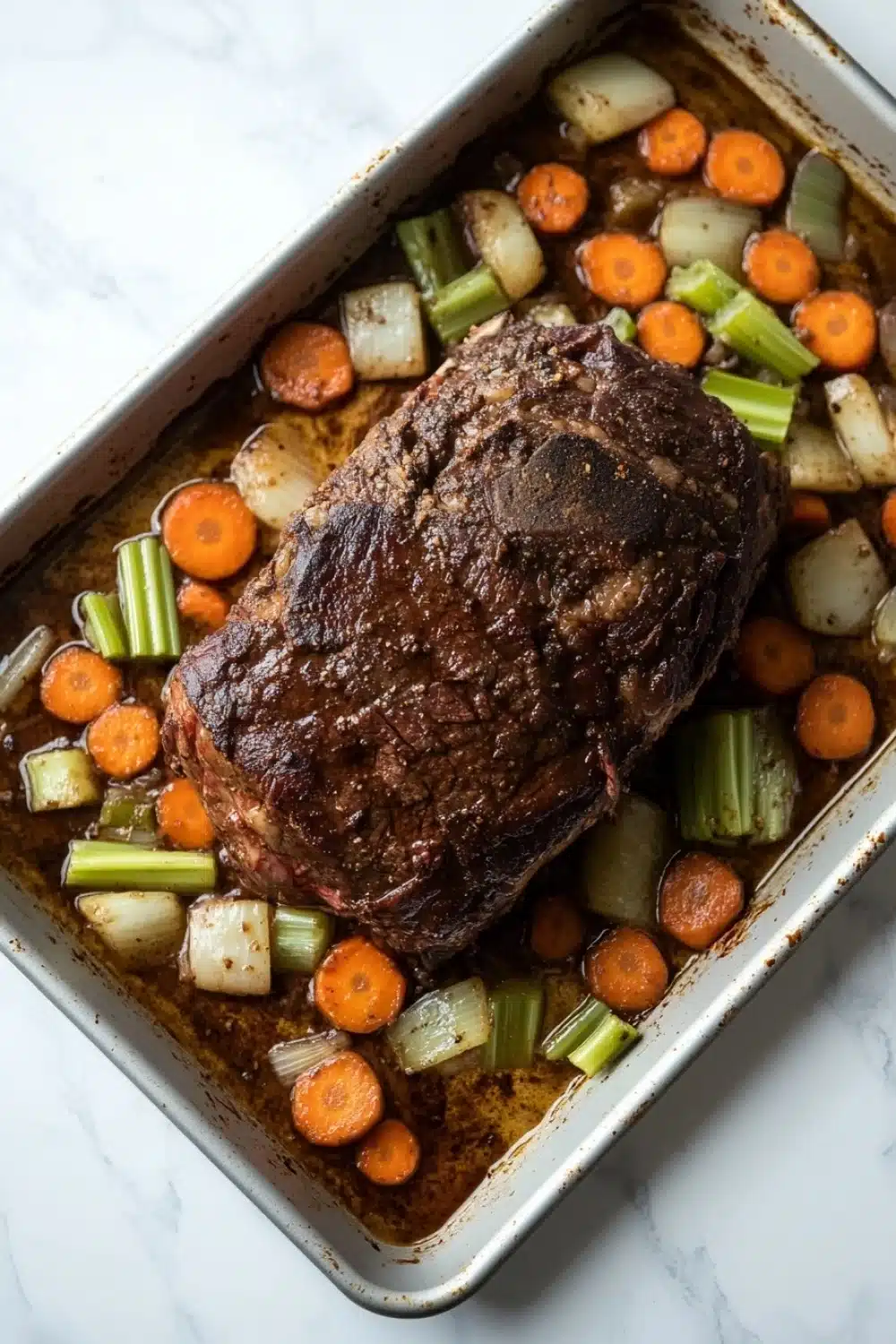 Slow cooker roast beef from frozen - the image shows a rectangular baking dish with a large piece of cooked meat in it. the meat appears to be a roast beef, with a dark brown crust on top. it is surrounded by a bed of diced carrots, celery, and onions. the dish is sitting on a white marble countertop. the vegetables are arranged in a way that they are overlapping each other, creating a colorful and appetizing display.