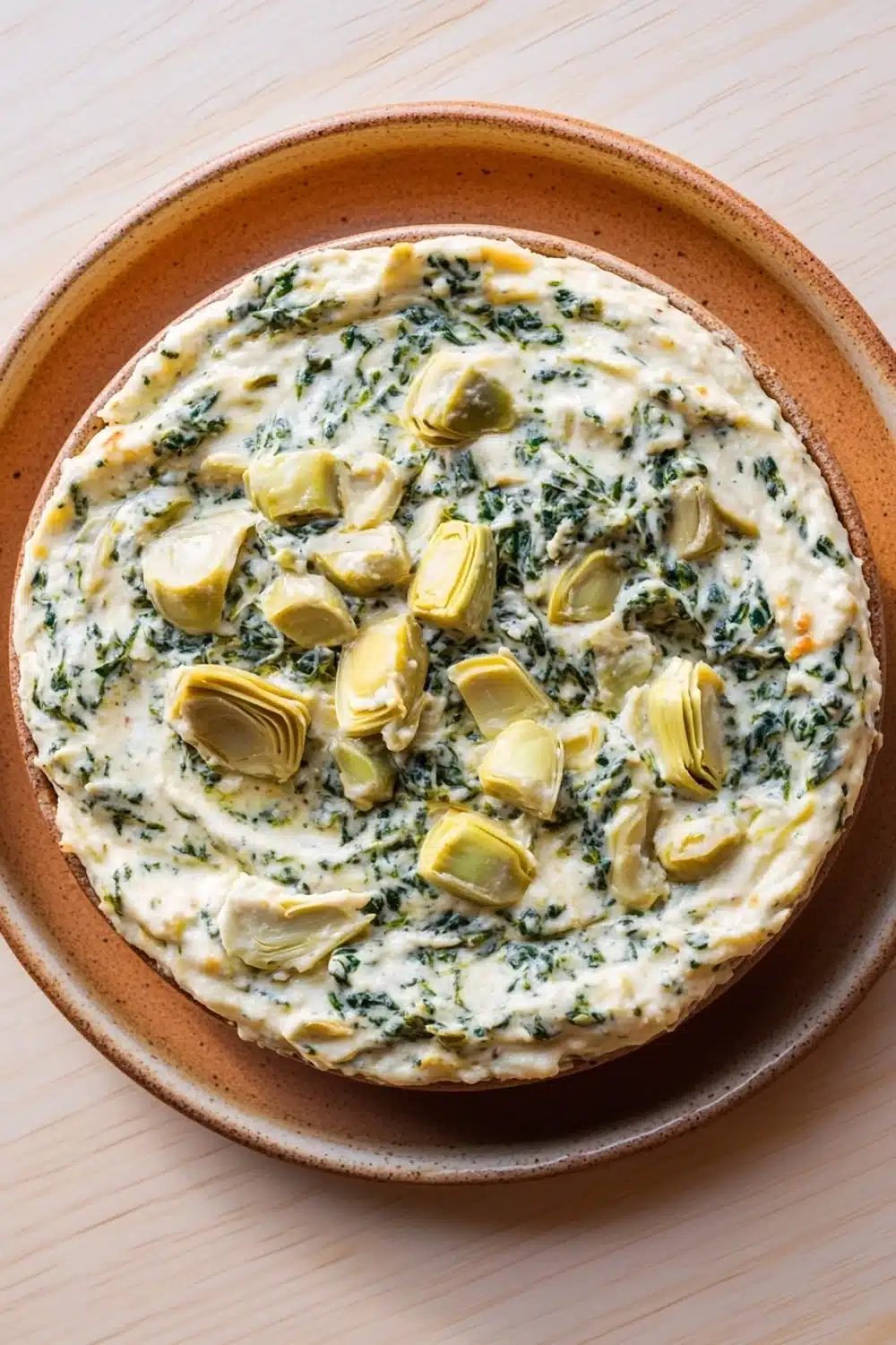 Spinach artichoke dip no cheese - the image is a close-up of a round pizza in a brown ceramic dish. the pizza is topped with a creamy white sauce and garnished with artichokes and spinach. the sauce appears to be thick and creamy, and the artichoke pieces are arranged in a circular pattern on top of the sauce. the dish is sitting on a wooden table.