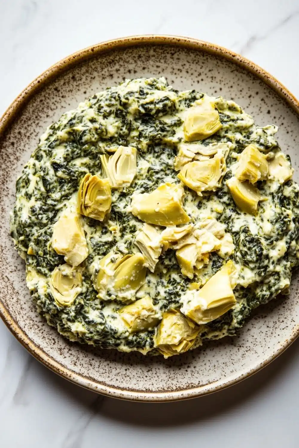 Spinach artichoke dip no cheese - the image is a close-up of a plate of food. the plate is round and has a brown rim with a speckled pattern. on top of the plate, there is a creamy green sauce with artichokes scattered throughout. the sauce appears to be a type of spinach or spinach-based dish. the artichoke pieces are cut into small cubes and are arranged in a circular pattern on the plate. the dish is sitting on a white marble countertop.