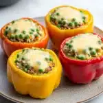 Stuffed bell peppers kids - the image shows four stuffed bell peppers on a white marble plate. the bell peppers are in different colors - orange, yellow, red, and green. each bell pepper is filled with a mixture of ground beef, rice, and peas, and topped with melted cheese. the plate is garnished with a sprig of fresh green herbs. the background is blurred, but it appears to be a kitchen countertop.