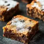 Sugar cookie brownies - the image shows a close-up of a freshly baked chocolate chip cookie bars on a black baking tray. the bars are golden brown in color and have a crumbly texture. they are dusted with powdered sugar, giving them a shiny appearance. the chocolate chips are visible on top of the bars, adding a pop of color to the dish. the tray is lined with parchment paper, and the background is blurred, making the bars the focal point of the image.