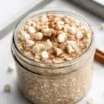 Sugar cookie overnight oats - the image shows a glass jar filled with a creamy, light-colored dessert. the dessert appears to be made with oats, nuts, and marshmallows, and is topped with a sprinkle of cinnamon. the jar is sitting on a white marble countertop with a few cinnamon sticks scattered around it. the background is blurred, but it seems to be a kitchen countertop.